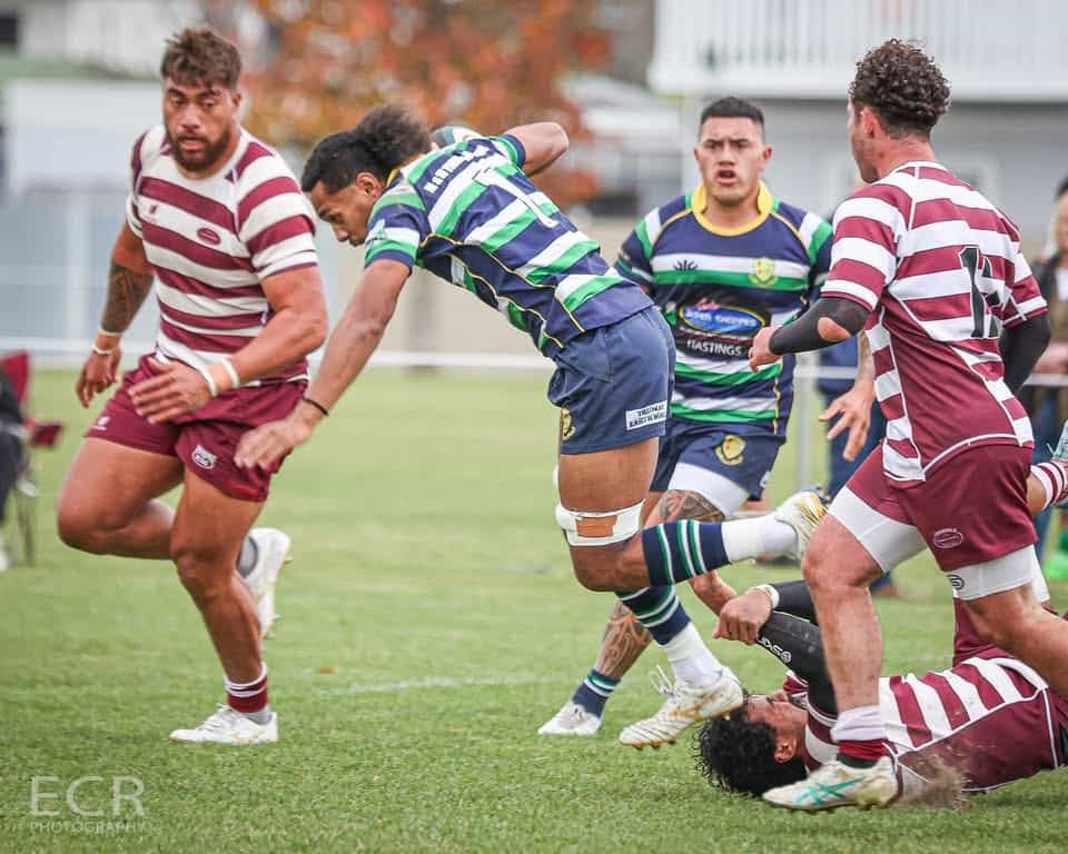 A rugby player in a green and blue jersey runs with the ball, stepping past defenders in maroon and white jerseys